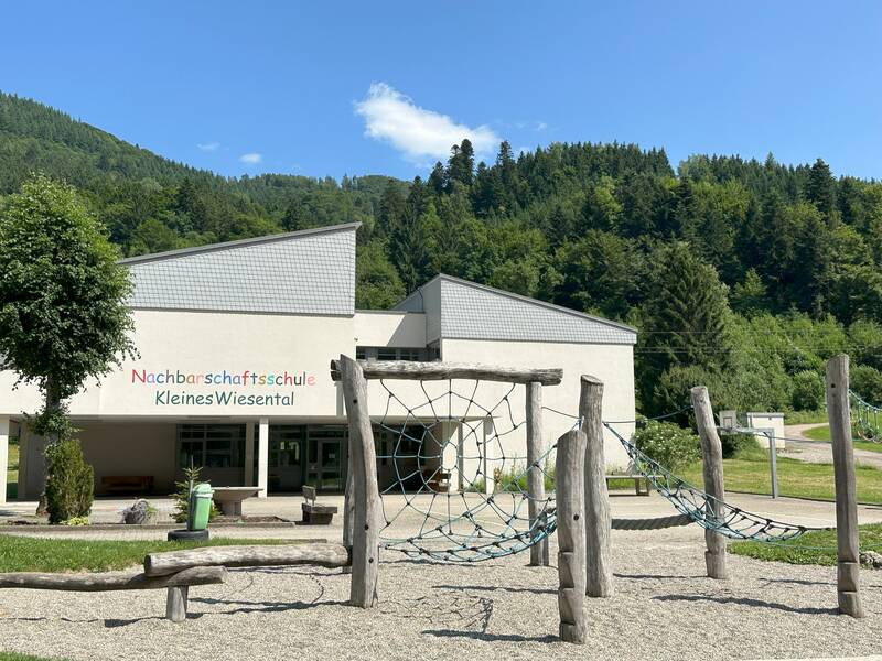 Vor einem Hintergrund aus grünen Hügeln steht die "Nachbarschaftsschule Kleines Wiesental", mit einem Klettergerüst aus Holz und Seilen im Vordergrund.