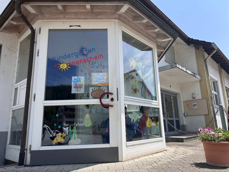 Der Eingang eines Kindergartens mit der Aufschrift "Kindergarten Sonnenschein" auf der verglasten Tür, die mit bunten Papierfischen dekoriert ist.