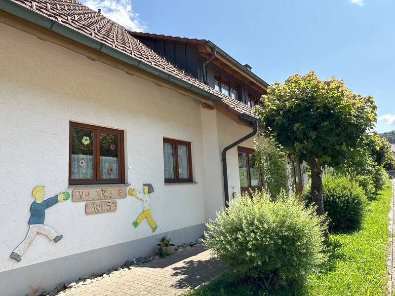 Ein einladendes, helles Gebäude mit einer dekorativen Wandbemalung von spielenden Kindern und gepflegter Begrünung an der Seite.