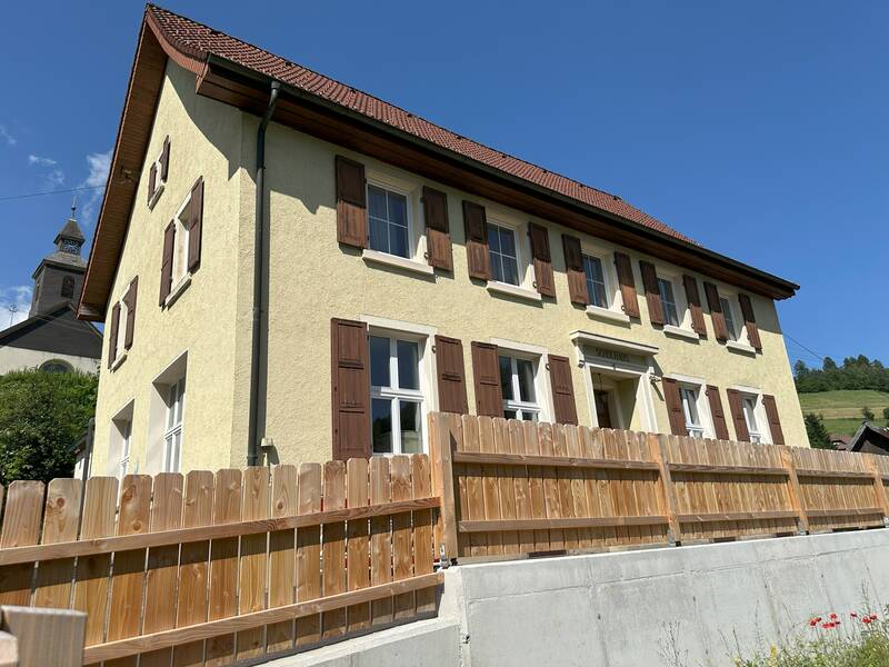 Ein zweistöckiges hellgelbes Haus mit braunen Fensterläden steht hinter einem Holzzaun und einem Betonfundament vor einem blauen Himmel.