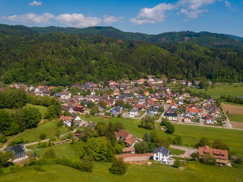 Ein kleines Dorf mit bunten Häusern liegt eingebettet in einer grünen, bewaldeten Hügellandschaft unter einem blauen Himmel mit weißen Wolken.