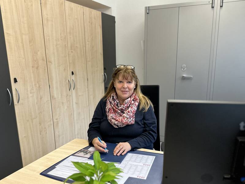 Eine Frau sitzt lächelnd an einem Schreibtisch in einem Büro und hält einen Stift in der Hand, umgeben von einem Kalender und einer Zimmerpflanze.